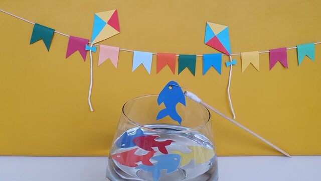 Kites, flags, colorful fish in the jar with water, fishing rod, on the table. Yellow background. Tradition to brighten up the Brazilian June party is the play and fun of toy fishing. Space for text.