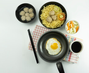 Indonesian Warm noodles served with eggs and fresh tomatoes with meatballs on a black bowl and pickled on a white bowl and eggs on a white background