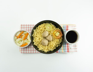 Indonesian Warm noodles served with fresh tomatoes and meatballs in a round bowl on black and pickles on a white bowl on a white background