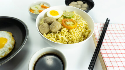 Indonesian Warm noodles served with fresh tomatoes and meatballs on a white round bowl and pickles on a white bowl on a white background