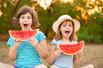 Two preschool smiling child sister sit with crossed legs on grass and eating watermelon at summer park with sunset light