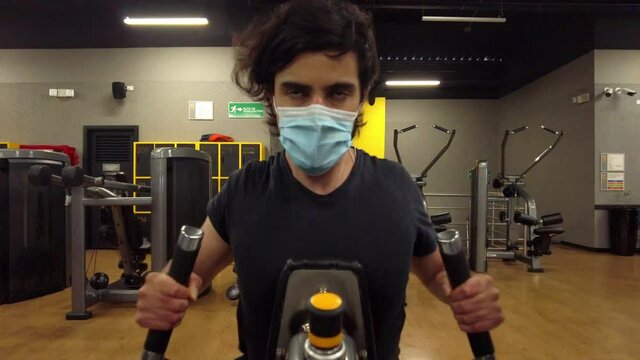 Young man training arms gym workout in a rowing machine using a medical face mask and black shirt during covid-19 or coronavirus pandemic. Dark background with yellow details and machines wood on flat