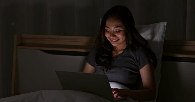 Asian cute woman laying on bed at night in bedroom and using notebook computer laptop with happy face and comfortable gesture.
