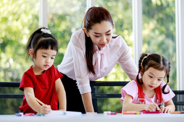 Portrait closeup shot of Asian beautiful female elementary school tutor teacher standing look after watching two little preschool girls drawing painting writing cartoon on paper book on table at home