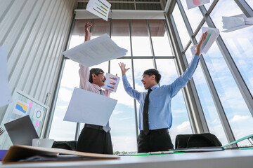 Two Asian professional businessmen celebrating their successful project by throwing document papers in the air with funny happy smiles in the daytime. Concept of achievement of a new start-up company