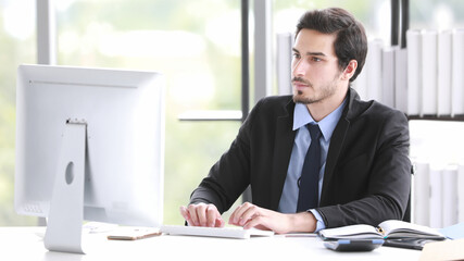 Good looking young adult businessman in suit sitting at desk and working with desktop computer in modern office