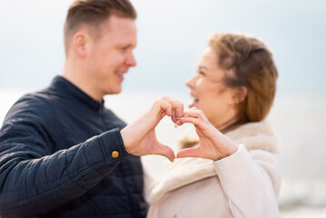 Two young couple is making heart, looking at each other and smiling while standing at the beach.
