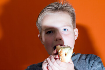 Handsome 10 yers old boy holding and biting hot dog closeup indoors orange studio background image.Close up.Front view.