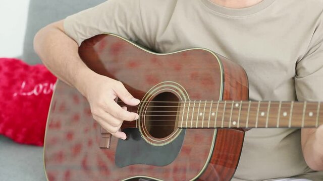 Slow motion and close up view of acoustic guitar being played by a man wearing bedge color t-shirt on sofa at home