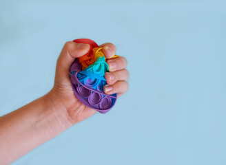 A child's hand squeezes a silicone colorful toy Pop It. Solid blue background. 