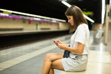 Young positive woman using mobile phone sitting at underground platform