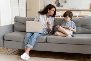 Fototapeta na wymiar Curious mother look at tablet screen while little preschool boy play games on digital pc. Young remote working mom with small kid at home at lockdown. Freelance businesswoman mum with laptop on couch