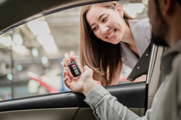 Car salesman with smiling face sending the car key to customer. Happy Asian woman giving key to customer for buy a new car or vehicle rental in automobile showroom.