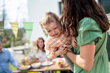 Crying baby with mother outdoors in garden at home, birthday celebration party.
