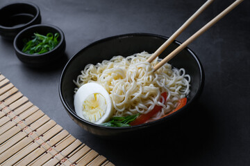 Black bowl with noodle, egg and vegetables and chopsticks near at black background.