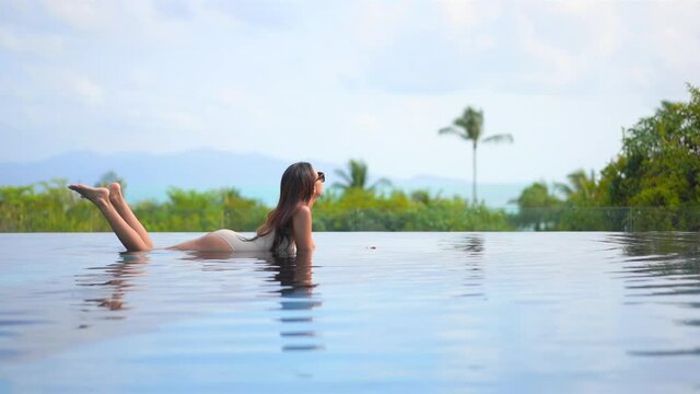 Sexy asian woman lying in shallow water of infinity pool with breathtaking view on tropical vegetation, static slow motion