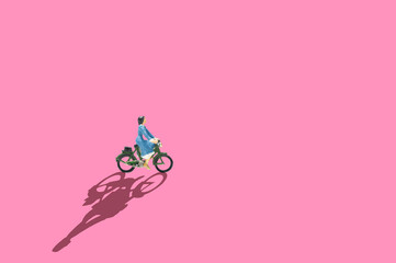 Fototapeta na wymiar Selective Focus miniature woman blue dress ride bicycle on pink background and long shadow.