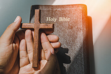 hand with wood cross over the bible on wood table with window light.christian backgound
