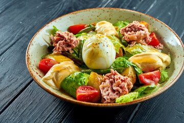 Traditional salad of French cuisine - nicoise with tuna, asparagus, tomatoes, potatoes and poached egg served in a bowl on a black wooden background