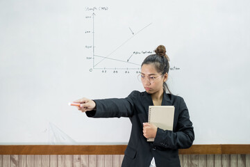An Asian female teacher in a classroom near the whiteboard is writing and teaching a lesson with a...