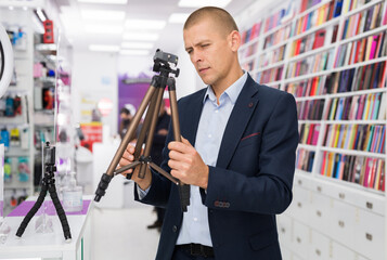 Elegant young man standing in tech store with tripod for camera in his hands