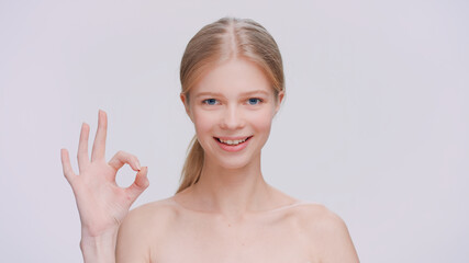 Young blonde woman makes an ok sign, looks at the camera and smiles. 