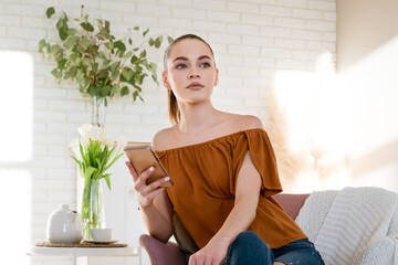 young beautiful woman of European appearance in casual clothes sits in an armchair at home in bright room with phone in her hand with pensive look. Nearby is table with teapot and cup vase of flowers