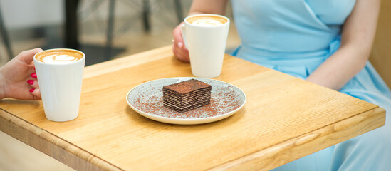 Two young women with cups of coffee and pieces of cake sitting at the table in a cafe outdoors