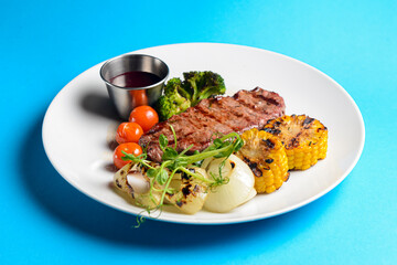 Delicious grilled beef steak with bbq vegetables and corn served on a white plate with sauce over blue background.