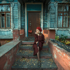 Woman in a burgundy coat and beret is patiently waiting sitting on the porch of an old wooden house.