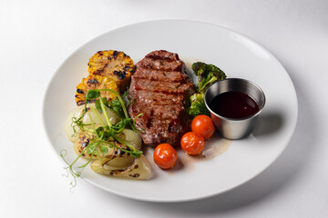 Delicious grilled beef steak with bbq vegetables and corn served on a white plate with sauce over white background.