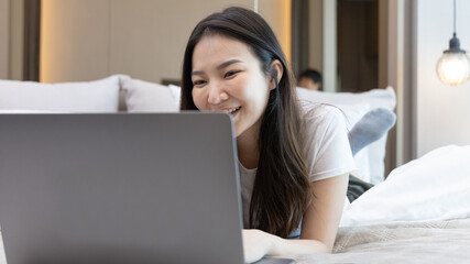 Asian woman uses laptop on bed in modern bedroom at home after she wakes up in the morning, Entertaining or relaxing using laptop technology, Wake-up activities, Happy lifestyle.