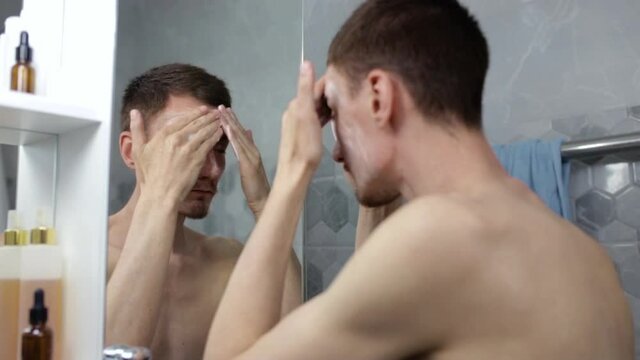 Handsome man applying skin care facial mask and looking in mirror in bathroom during daily beauty routine 
