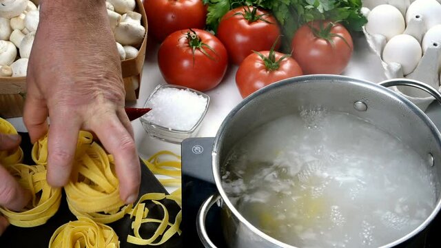 Chef puts dry pasta in saucepan with boiling water. Preparation of italian tagliatelle in domestic kitchen. Hands putting pasta to saucepan. Different vegetables and eggs on table. Close-up.