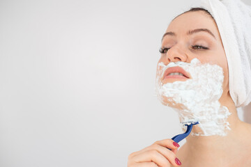 A woman with a white terry towel on her head and with shaving foam on her face shaves on a white...