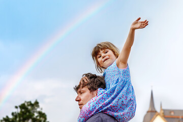 Little preschool girl sitting on shoulder of father. Happy toddler child and man observing rainbow on sky after summer rain. Happy family, bonding, love. Summertime. Dad and daughter. Father's day.