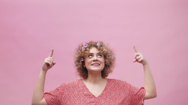 Happy funny young woman with flowers in curly hair pointing fingers above her head. Isolated on pink background