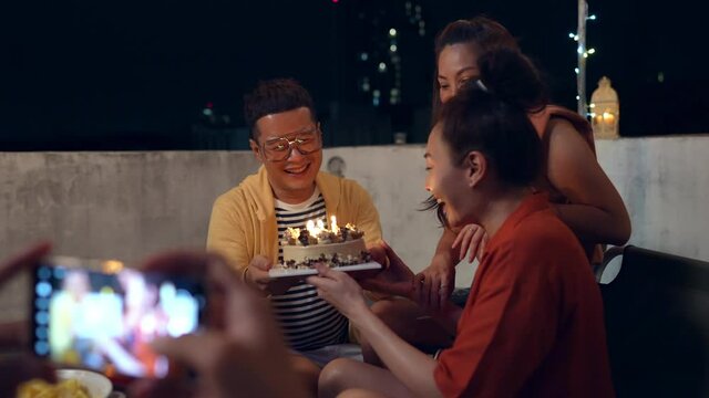 Asian man using smartphone taking photo or home video while celebration outdoor birthday party to female friend together. Woman excited with birthday cake and blowing birthday candle with happiness.