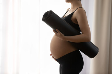 Close up cropped profile of pregnant woman in black sportswear holding yoga mat, touching belly,...