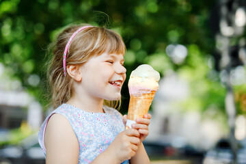 Little preschool girl eating ice cream in waffle cone on sunny summer day. Happy toddler child eat...