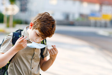 Happy kid boy with glasses and medical mask due to corona virus covid pandemic. Schoolkid with satchel waiting for bus on the way to school on sunny day. Healthy child outdoors on the street.
