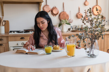 young asian girl in silk pajamas reading book while having breakfast in kitchen