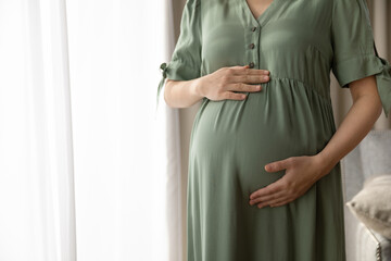 Close up cropped of pregnant woman in dress touching caressing belly, feeling baby kicks, standing at home, young future mother expecting child, enjoying healthy pregnancy, motherhood concept