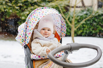 Cute little beautiful baby girl sitting in the pram or stroller on cold snowy winter day. Happy smiling child in warm clothes, fashion stylish baby coat. Baby's first snow. Winter walk outdoors.