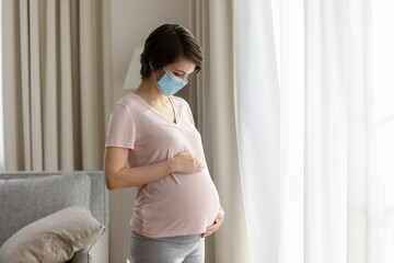 Pregnant woman wearing medical face mask standing, touching caressing belly, worried about childbirth during pandemic, covid-19, young future mom feeling anxiety, pregnancy and healthcare concept