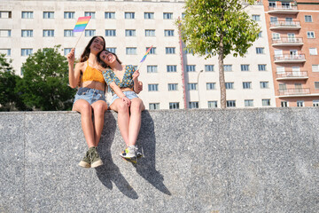 Lesbian couple holding flags, hugging and laughing sitting on a wall at street.