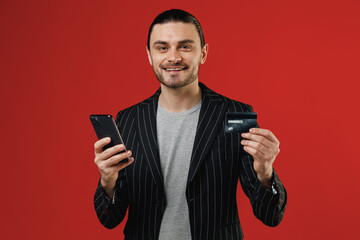 Young businessman latin man in black striped jacket grey shirt using mobile cell phone hold credit bank card doing online shopping order delivery booking tour look camera isolated on red background