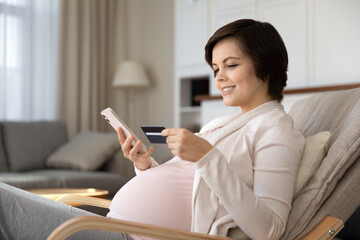 Smiling pregnant woman paying online by credit card, using phone, making secure internet payment, satisfied customer happy young future mom shopping in internet at home, browsing banking service - Powered by Adobe