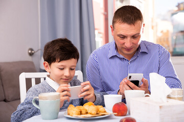 Fototapeta na wymiar Portrait of young man and his son using phones at table with tea indoors