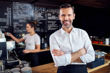 Portrait of smiling cafe manager standing in coffee shop with employee in background